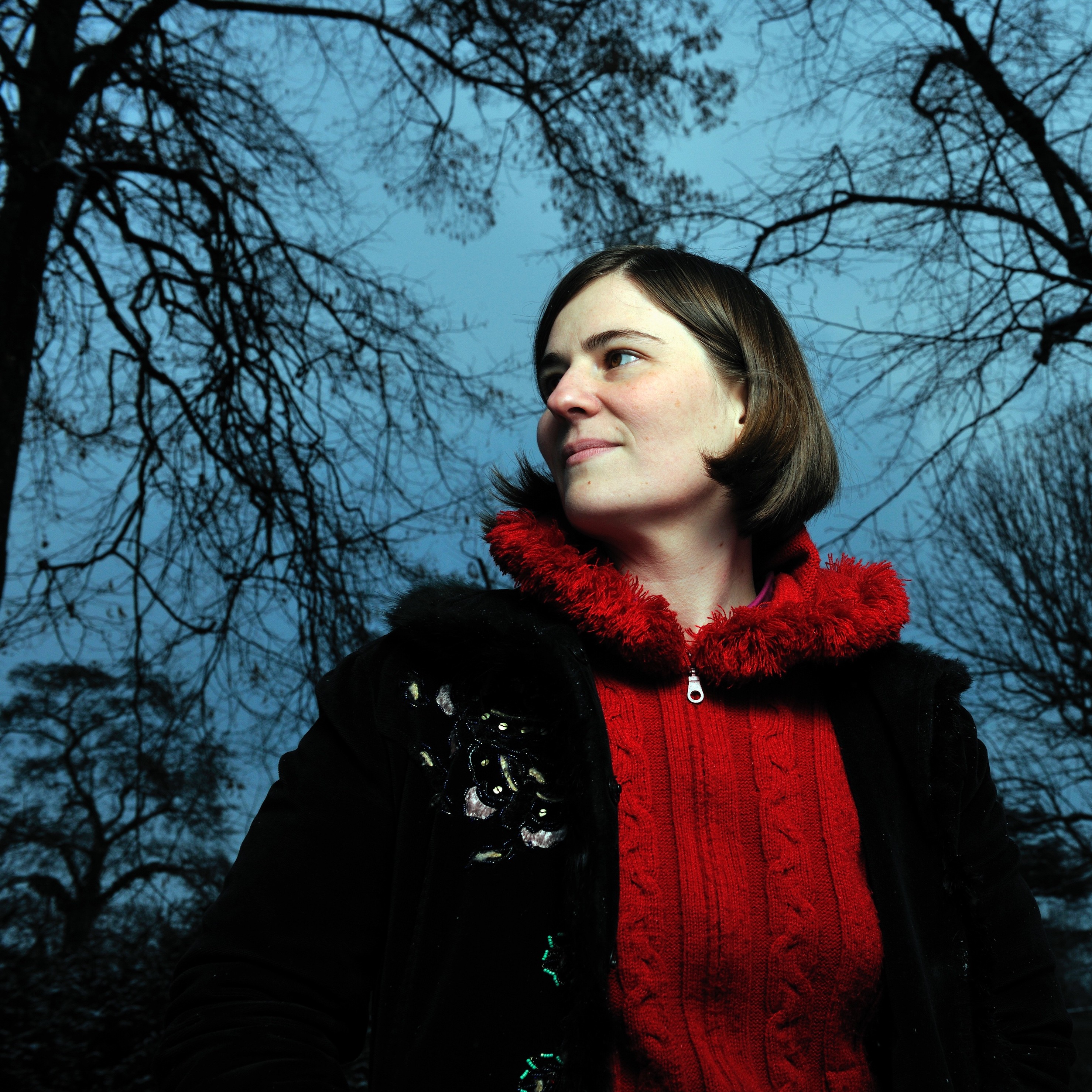 Portrait de Marie-Jeanne Urech. Cheveux bruns au carré, elle pose sur son profil droit gauche. En arrière plan, on voit les branches d'un arbre dénudé au-dessus d'elle et sur les côtés. Elle porte un pull rouge à fermeture éclaire, avec col montant. Il contraste avec sa veste noire, ouverte.